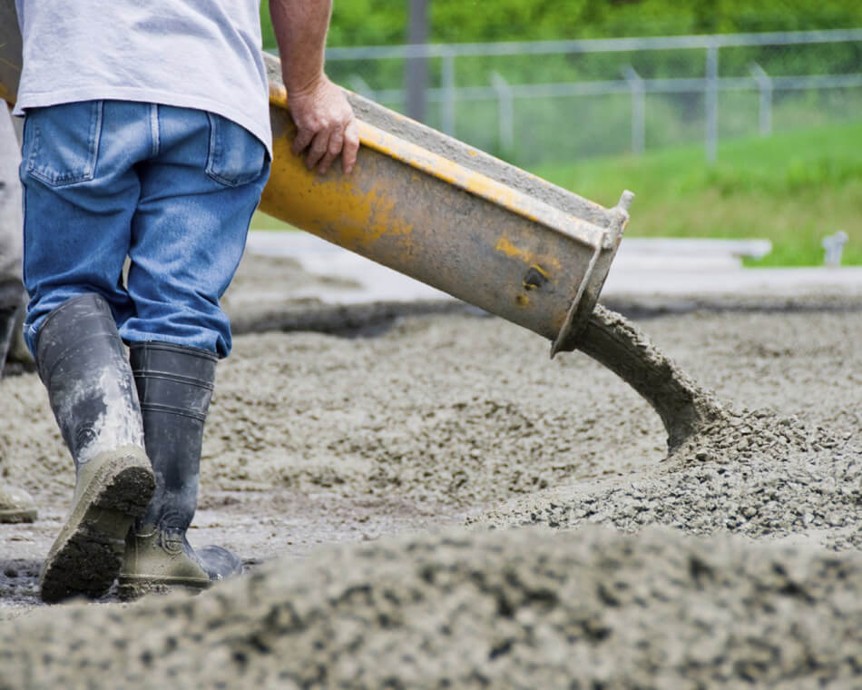 Construction worker in boots and jeans guides a concrete chute pouring wet concrete onto a gravel surface.